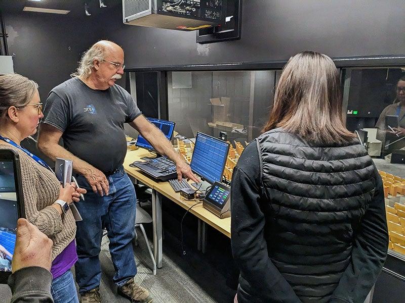 three people stand in a control booth with computers - a window looks out to auditorium seating below