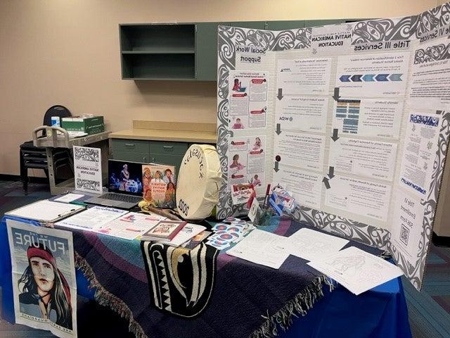 Table with a trifold chart displaying information, a native drum, books, and a laptop showing a Native performance